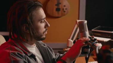 Kafkasyalı erkek müzisyenin, kayıt stüdyosunda ekipman kurarken, çalışma oturumuna hazırlanırken, mikrofonu ayarlarken, masanın üzerinde sentezleyici ile orta ölçekli yakın çekimi.