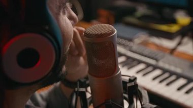 Genç erkek müzik yapımcısının kulaklıklarıyla stüdyoda, sentezleyici ve bilgisayarın önünde otururken, podcast 'e katılırken mikrofona konuşurken aşırı yakın çekim görüntüsü.