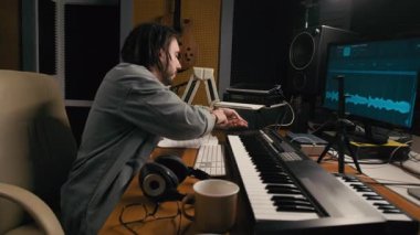 Beyaz tenli erkek müzisyenin kayıt stüdyosunda bilgisayar ekranlarının önünde otururken, sentezleyiciyi fişe takarken, yeni müzik üzerinde çalışmadan önce sesi kontrol etmek için akorlar çalarken orta ölçekli bir fotoğrafı.