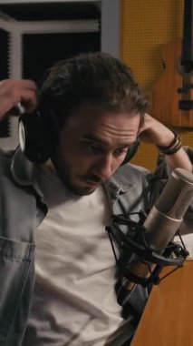 Genç müzisyen ya da podcast sunucusunun orta boy dikey yavaş portresi, kayıt stüdyosundaki mikrofonun önündeki sandalyede oturuyor, kameraya bakarken kulaklıkları çıkarıyor.