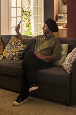 Şapka takan genç bir adam, rahat bir şekilde koltukta otururken rahat bir şekilde rahat bir oturma odasında selfie çekerken çağdaş dekorun bir karışımını sunuyor.