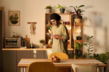 Kadın, çeşitli bitkiler ve ahşap raflarla dolu rahat bir mutfakta yemek hazırlıyor. Ilık aydınlatma davetkar bir atmosfer yaratır, dingin ortamı güçlendirir.