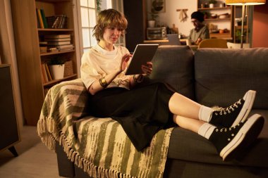 Kanepede oturup tablet okuyan bir kadın. Kitap rafları ve arka planda çalışan bir kişi ile rahat oturma odası ayarları