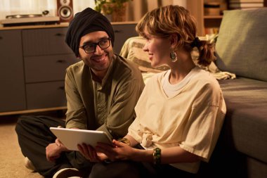 Gülümseyen çift sıcak oturma odasında oturmuş tabletleri birlikte tutuyorlar. Birbirine eşlik eden ve teknolojiyle eğlenen, neşeli bir anı paylaşıyorlar.