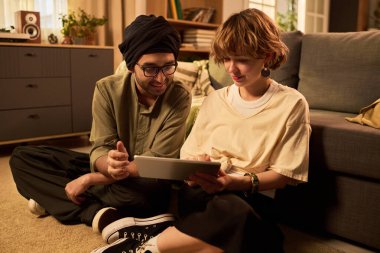 Çift, oturma odasında tablet kullanarak kaliteli zaman geçirmenin keyfini çıkarıyor. Raflar ve kanepe ile samimi ortam bağlanma ve sohbet için rahat bir ortam yaratıyor.