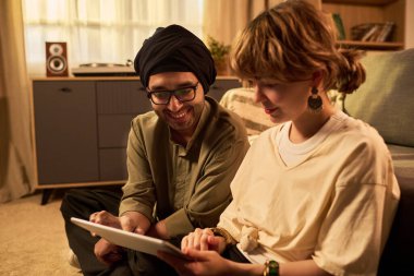 İki kişi sıcak bir odada oturmuş, bir tablet cihazıyla uğraşıyorlar. Odada sıcak bir ışık ve rahat bir atmosfer var.