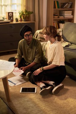 Genç bir adam ve kadın, oturma odasında, rahat bir oturma odası ve kitap rafları olan tabletin yanındaki belgeleri incelerken sohbet ediyorlar.