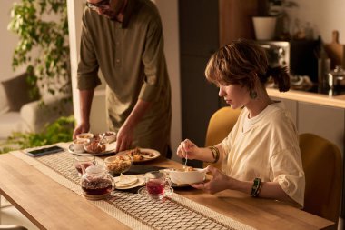 Genç kadın ve yaşlı adam modern mutfak masasında kahvaltıdan zevk alıyor, gündelik sohbetler yapıyor ve yemek seçimini takdir ediyorlar. Çeşitli yemek ve içecekler içeren masa ayarları