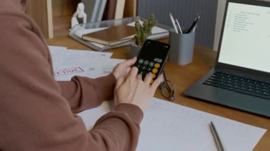 Akıllı telefon uygulamasındaki iç harcamaları hesaplayan, sonra da dizüstü bilgisayarın önünde bütçe planlayıcısı ile birlikte otururken fatura numaralarını yuvarlak içine alan, tanınmayan genç kadın ev sahibinin orta boy fotoğrafı.