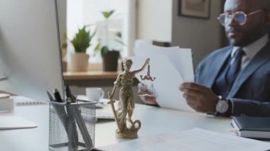 Hukuk bürosundaki Bayan Adalet Heykeli 'nin orta ölçekli bir resmi. Gözlüklü ve takım elbiseli genç bir Afro-Amerikan erkek avukat. Bulanık arka planda oturup yasal belgeleri inceliyor.