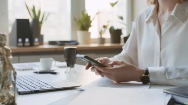 Beyaz gömlekli kadın avukat ya da hukuk firmasında ya da noter ofisinde masasında oturmuş müşteriyle iletişim kurarken akıllı telefondan mesajlar yazarken orta ölçekli orta ölçekli bir fotoğraf.