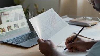 Tanımlanamayan Afro-Amerikan bir adamın evde masasında oturması, gecikmiş faturaları tutması, kamu hizmetleri ödemelerini hesaplaması, deftere numaralar yazması, kişisel bütçesini takip etmesi.