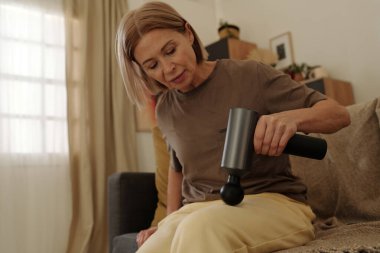 Ahşap dekorlu ve hafif ışıklandırmalı rahat oturma odasında saç kurutma makinesi kullanan son sınıf öğrencisi bir kadın. İşine odaklan, günlük rutine ve kişisel bakıma bir göz at.