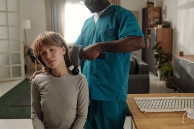 Genç insan koltukta otururken mavi önlüklü profesyonel saçlarını kurutuyor, rahat, aydınlık bir odada yumuşak mobilyalar ve bitkilerle.