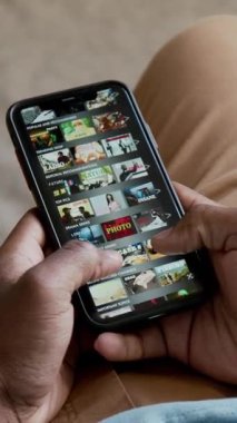 Tanımlanamayan Afro-Amerikan bir adamın ellerini dikey çekerek akıllı telefondan online sinema uygulamasını izlemek ve evde tek başına dinlenmek için film listesine kayıyor.