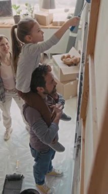 Dikey çekimde, genç bir adamın omuzlarında oturan ve boyacı ile oturma odasının duvarını boyarken ailesine yeni dairelerini yenilemeleri için yardım eden sevimli küçük kız görülüyor.