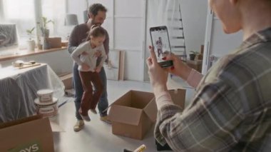 Genç baba, yeni dairenin oturma odasındaki karton kutuya küçük şirin bir kız yerleştirirken bir kadın önlerinde duruyor ve akıllı telefondan video kaydediyor.