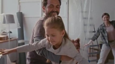 Mutlu genç sakallı adam kameranın önünde yeni evlerinin oturma odasında dans eden heyecanlı bir kadına karşı oynarken sevimli, gülümseyen küçük kızıyla oynuyor.