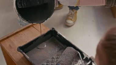 Plastik kovayı tutan ve tepsiye gri boya döken genç bir kadının eldivenli elini eğ. Küçük kızının yanında durup, evi yenilemesine yardım et.