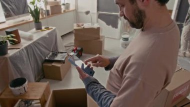 Genç adam oturma odasında kameranın önünde dururken cep telefonu ekranını işaret ederek ev yenileme fikirleri hakkında çevrimiçi bilgi ya da video ararken ya da mesajlaşırken