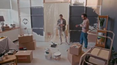 Genç bir adam ve kadının yeni dairelerinin oturma odasında tadilat çalışmaları, duvarları boyamaları, paketlenmiş karton kutular taşımaları ve ev eşyalarını onarmaları.