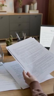 Evdeki masasında oturan, dizüstü bilgisayar ekranı ve kağıt kopya üzerinde ipotek sözleşmesi taslağını inceleyen ve yeni bir ev almayı planlayan isimsiz bir kadının elleri dikey olarak yakın plan.