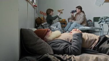 Mülteci sığınağında yatağın üzerinde uyumaya çalışan, iç çeken, oynayan, oynayan, çay içen, annesi olan, gri sakallı, yaşlı, beyaz bir adamın orta boy fotoğrafı.