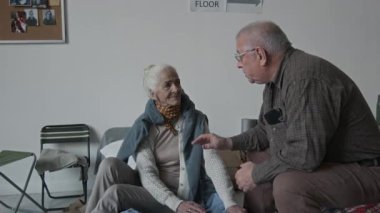 Orta boy beyaz bir adam, geçici bir acil durum barınağında, katlanır yatakta oturuyor, yaşlı bir bayanı teselli ediyor, doğal afetten kaçıyor, konuşuyor ve omzunu okşuyor.