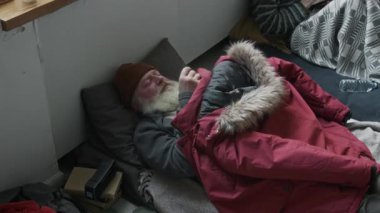 Mülteci kampında şiltede yatan gri sakallı, orta yaşlı beyaz bir adam, uyumaya çalışıyor, yün şapkasını gözlerine çekiyor ve üstünü parka ceketiyle örtüyor.