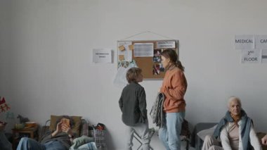 Orta ölçekli, kaygılı, beyaz anne ve oğlu mülteci barınağındaki ilan panosundaki kayıp şahıs fotoğraflarına bakıp derme çatma yatağa yürüyor, oturuyor, sarılıyor, battaniyeye sarılıyor.