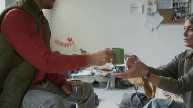 Doğal afet sırasında mülteci kampında birlikte kalırken olgun bir kadınla sıcak çay içen, orta boy beyaz bir erkek resmi.