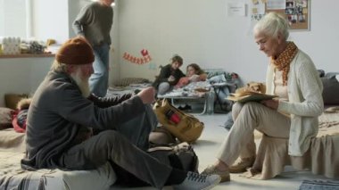 Mülteci barınağında kalan üst düzey beyaz bir çift, koca çantada arıyor ve kitap okuyan karısına kazak veriyor, onlar da ona teşekkür ediyor ve omuzlarını sarıyor.