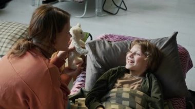 Mülteci kampında yatağın üzerine uzanmış, 8 yaşında oyuncak ayıyla gülen neşeli, beyaz bir kadının doğal afetler sırasında çocuğu neşelendirdiği orta boy bir fotoğraf.