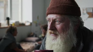Evsiz, gri sakallı, bereli, mülteci barınağında çay içen, savaştan sonra evinden ayrılan, endişeli ve üzgün bir şekilde kameraya bakan beyaz bir adamın yakın plan portresi.