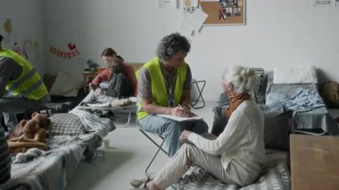 Sarı yelekli gönüllüler savaş ya da mülteci sığınaklarındaki felaket nedeniyle yerlerinden edilen insanlara yardım ediyor. Elinde dosya olan beyaz bir kadın sandalyeye oturmuş, yaşlı bayanı ihtiyaçları hakkında sorguluyor.