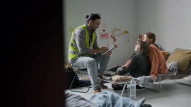 Mülteci barınağındaki kamp sandalyesinde oturan, anne ve çocukla konuşan, acil yardıma koşan, sarı yelekli, neşeli, beyaz bir erkek gönüllü.