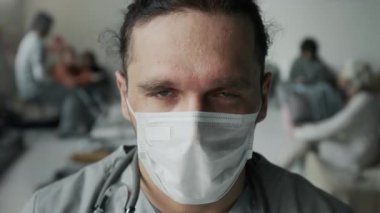 Beyaz maske takmış, kameraya bakıyor, savaştan ya da felaketten etkilenen mülteci barınağında yaşayanlara yardım ederken mavi üniformalı beyaz erkek hemşirenin yakın plan portresi.