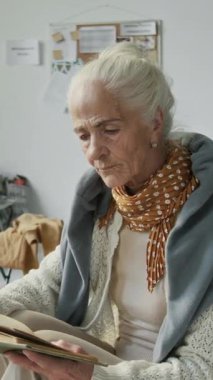 Mülteci kampında oturan, doğal afetten ya da savaştan etkilenmiş, kitap okumaktan zevk alan, kameraya bakıp gülümseyen mutlu, yaşlı beyaz kadının orta boy dikey portresi.
