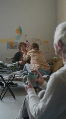 Mülteci barınağında, beyaz saçlı yaşlı bir kadının, doğal afetten ya da savaştan kaçtıktan sonra, arka planda oğluyla oynarken çekilmiş dikey resmi.