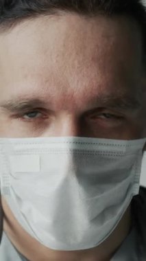 Tıbbi üniformalı, beyaz maske takmış, hastane veya mülteci barınağında çalışırken kameraya bakan beyaz erkek hemşirenin dik dik çekilmiş portresi.