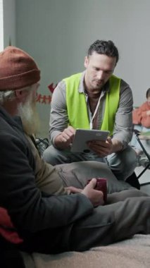 Sarı yelekli beyaz erkek acil durum görevlisinin tam dikey görüntüsü sakallı yaşlı erkek mültecinin yanında oturuyor, sorular soruyor, dijital tablet üzerindeki kontrol listesini dolduruyor.