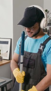 Çalışma kıyafetleri ve kulaklıklar takan neşeli, çoklu etnik bir adamın ofis çalışma alanını paspaslarken şarkı söylediği dikey çekim.