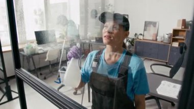 Beyaz üniformalı ve koruyucu eldivenli bir kadın cam duvarın yüzeyini dezenfekte ederken erkek meslektaşı ofis zeminini paspaslıyor.