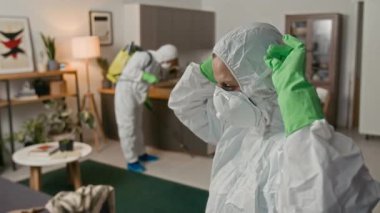 Tehlikeli madde giysisi içindeki kadın dezenfektanının odak noktası solunum cihazı ve gözlük takarken aynı zamanda arka plandaki erkek meslektaşla dezenfekte için hazırlanıyor.