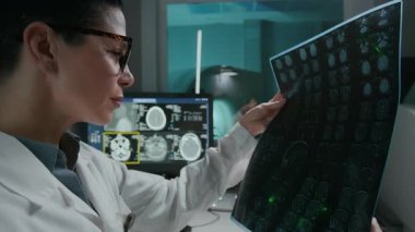 Orta yaşlı beyaz önlüklü beyaz kadın radyoloji uzmanı, gözlüklü, sinemada beyin MR 'ı inceleyen, dejeneratif hastalık belirtileri arayan orta büyüklükte bir kadın.