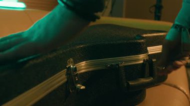 Siyah gitar kılıfını açan isimsiz müzisyenlerin yakın plan çekimleri ve loş ışıklı müzik aletleri.
