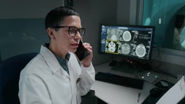 Kafkas kadın radyoloji uzmanı klinikte otururken, bilgisayar ekranında beyin MR sonuçlarını incelerken, hastayla mutlu haberleri paylaşmak için cep telefonuyla konuşurken orta boy çekilmiş bir fotoğraf.