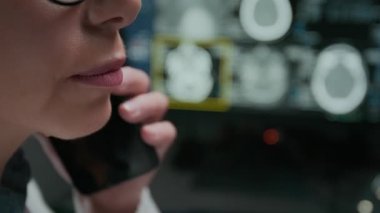 Cep telefonundan hastaya konuşan, beyin tomografisi ile ilgili rahatsız edici haberleri paylaşan tanınmayan kadın radyologların alt yüz ve ağızlarına aşırı yakın çekim, bulanık arka plandaki bilgisayarda MRI taraması