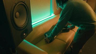 Yüksek açılı, günlük kıyafetlerle erkek müzisyenin gitar kılıfını kanepeye koyup, açıp loş neon ışıklı odada müzik aletini çıkardığı bir sahne.