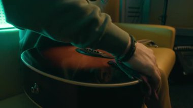 Loş neon ışıklı stüdyoda enstrüman çalmadan önce yüzsüz müzisyenin akustik gitarı bez parçasıyla temizlediği yakın çekim.
